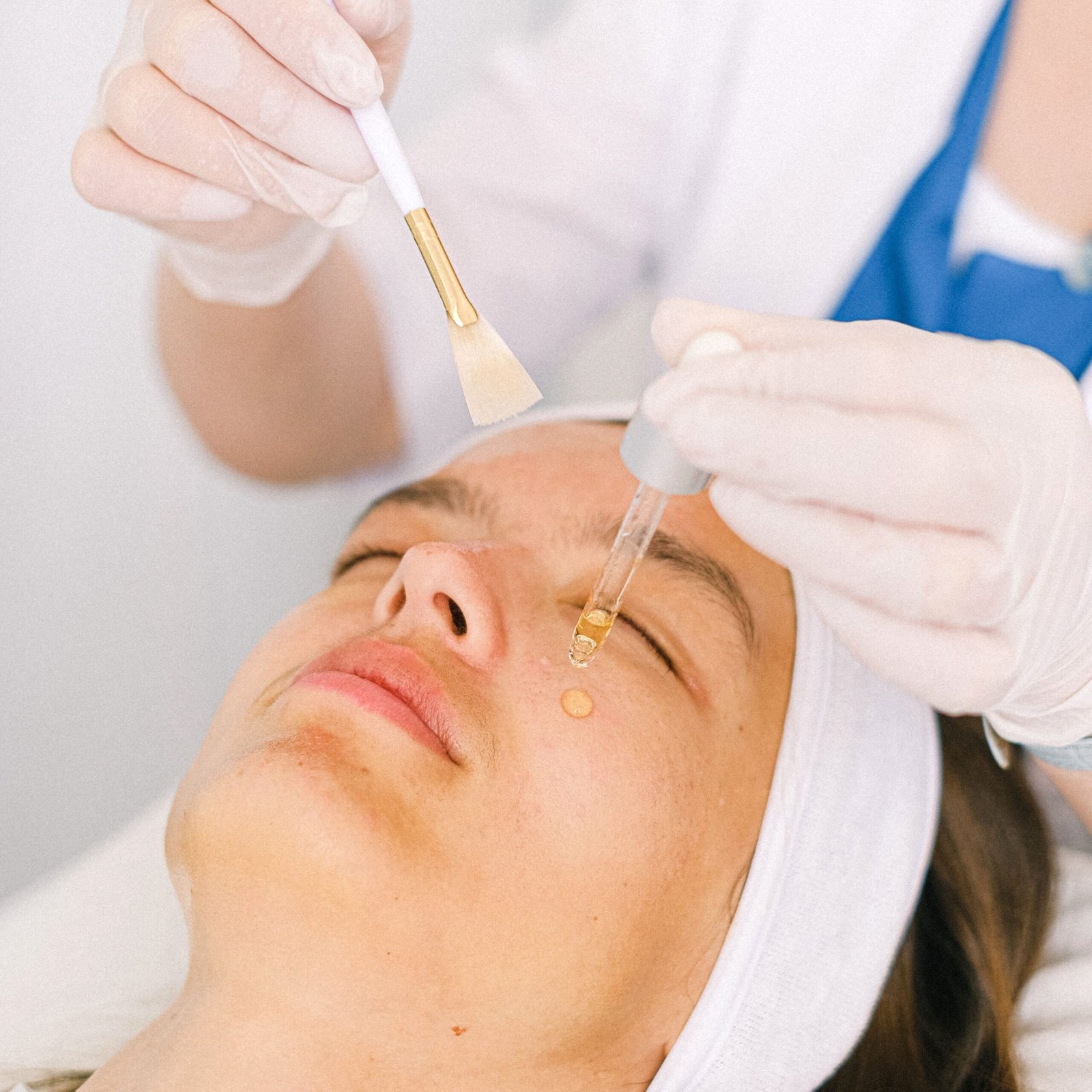 Skilled female beautician in uniform with brush applying moisturizing serum on beautiful female customer face skin in light modern beauty center